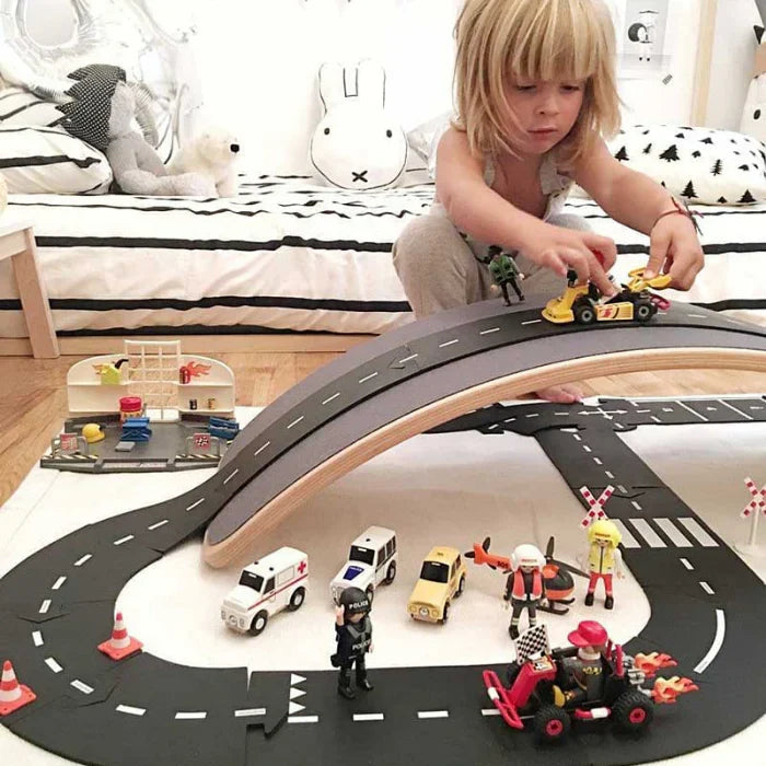 Child playing with a wooden toy road set with cars and figures on a bed.